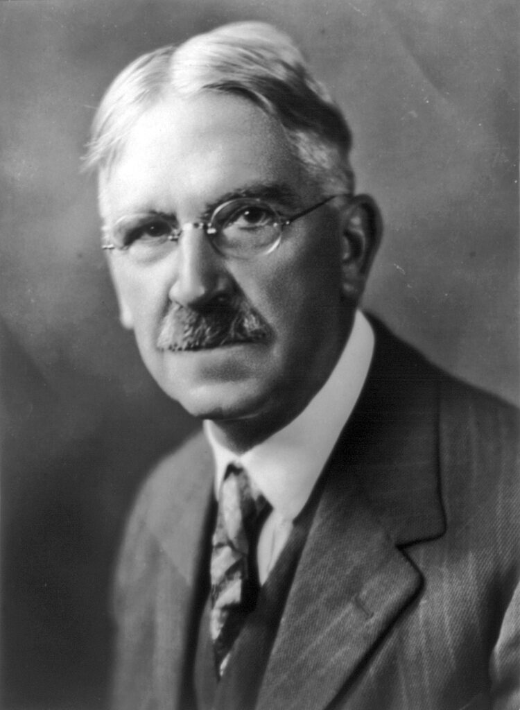 John Dewey A black and white photo shows an older man in a suit wearing thin wire frame glasses.