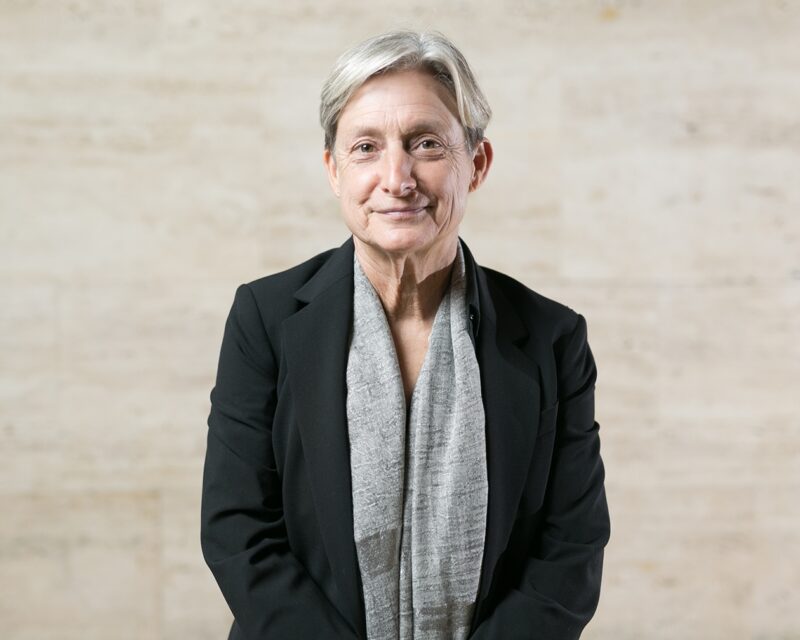 Judith Butler A person is shown from the waist up wearing a black blazer and a scarf.