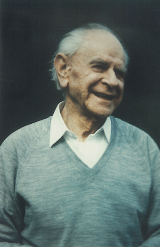 Karl Popper An older man is seen wearing a blue sweater with a white collared shirt under it. He is smiling with his head turned in a three-quarters view.