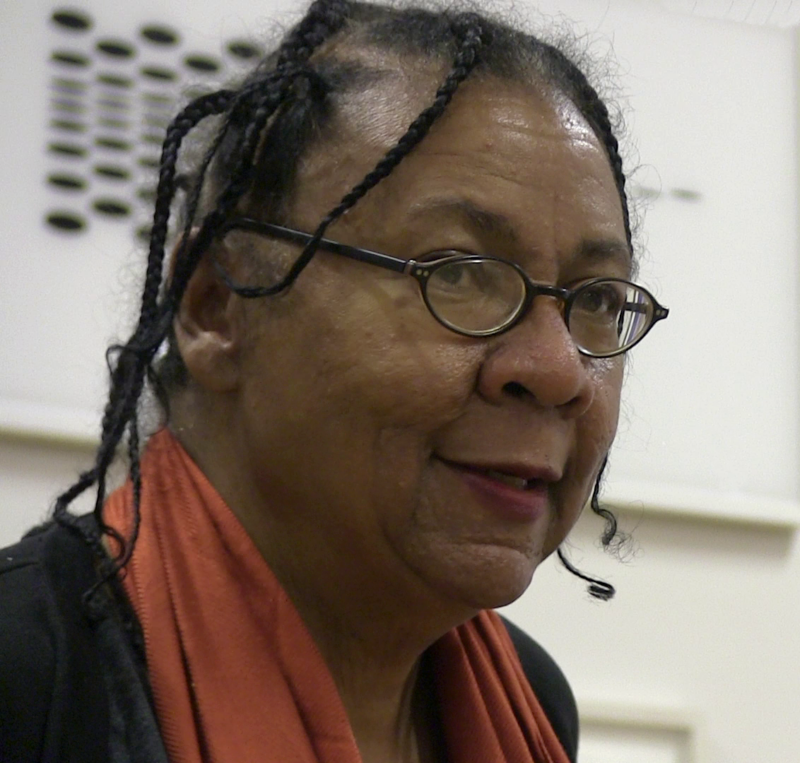 Bell Hooks A woman is shown in a color photo from the neck up. She is wearing a coral scarf and thin wire frame black glasses.